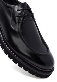 Zwarte leren schoen met een glanzende afwerking, ronde neus, decoratieve stiksels en een gestructureerde, dikke rubberen zool met uitgesproken looppatronen.