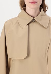 Trench coat beige con ampio colletto, presenta una chiusura a bottoni al collo e una texture liscia e opaca. Linee semplici e pulite dominano il design.