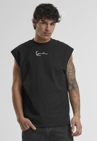 T-shirt en coton sans manches noir avec un logo signature blanc sur la poitrine. Le tissu a une texture lisse et une coupe décontractée.