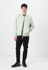 Lichtgroene bomberjack met een rits aan de voorkant, geribbelde manchetten en een subtiel logo. Gestreept shirt eronder, zwarte broek en sneakers.