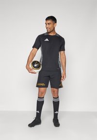 Mann in schwarzem Fußballtrikot, der einen schwarz-weißen Fußball hält, steht und schaut zur Seite vor einem schlichten Hintergrund.