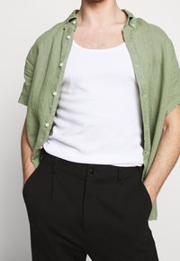 Vihreä pellavasta valmistettu lyhythihainen paita nappien kanssa, valkoisen topin päällä; yhdistetty mustiin housuihin. Materiaali vaikuttaa kevyeltä ja teksturoidulta.