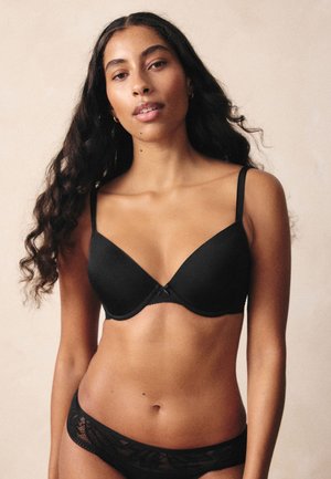 Žena s dugom kovrčavom kosom u crnom grudnjaku i odgovarajućem čipkanom donjem rublju, stoji na neutralnoj bež pozadini.