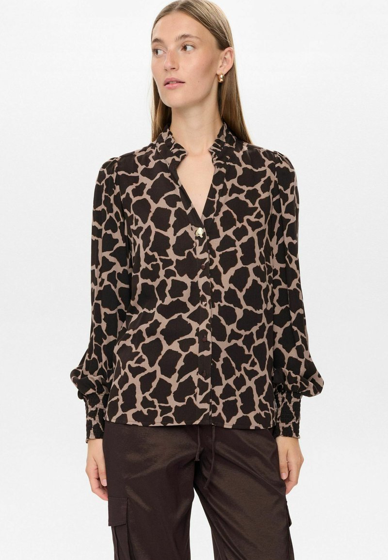 Donna che indossa una camicia a maniche lunghe con stampa giraffa marrone e nera e pantaloni cargo di satin marrone scuro, in piedi di fronte a uno sfondo bianco.