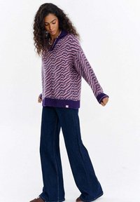 Kobieta o długich, kręconych włosach, ubrana w fioletowo-różowy sweter w wzory, szerokie ciemnoniebieskie jeansy i brązowe buty, stojąca z lekko rozstawionymi ramionami.