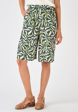Vrouw draagt groene shorts met een koordje en wit-gele bladpatronen, gecombineerd met bruine gewebde sandalen en een wit topje.
