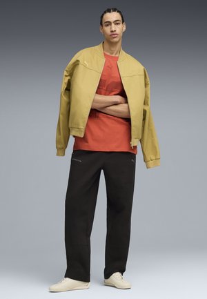 Homme aux cheveux tressés, bras croisés, portant une veste jaune sur un t-shirt rouge, pantalon noir et baskets blanches sur un fond gris.