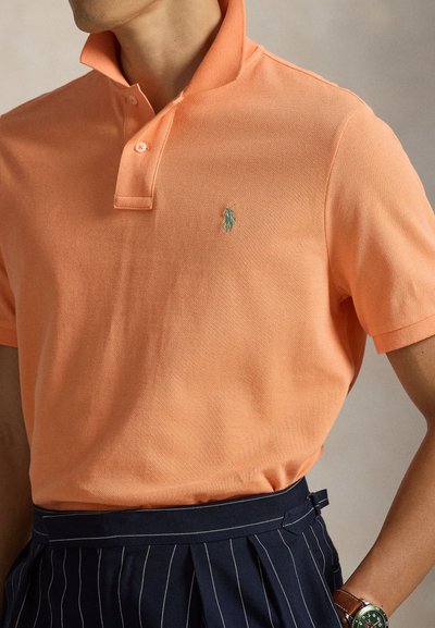 Πορτοκαλί πόλο μπλούζα με υφασμάτινη υφή, που διαθέτει ριμπ κολάρο και ένα μικρό πράσινο κεντητό λογότυπο στο στήθος. Συνδυάζεται με μια ζώνη με ριγέ λεπτομέρειες.
