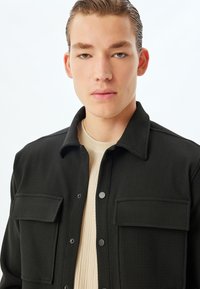 Giacca nera con texture, due tasche sul petto, bottoni a pressione e una morbida maglietta beige sotto. Linee pulite, design minimalista, vista frontale.