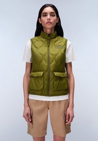Groene gewatteerde vest met hoge kraag, ritssluiting aan de voorkant en twee opgestikte zakken. Gepaard met een wit shirt en beige shorts. Gladde textuur.