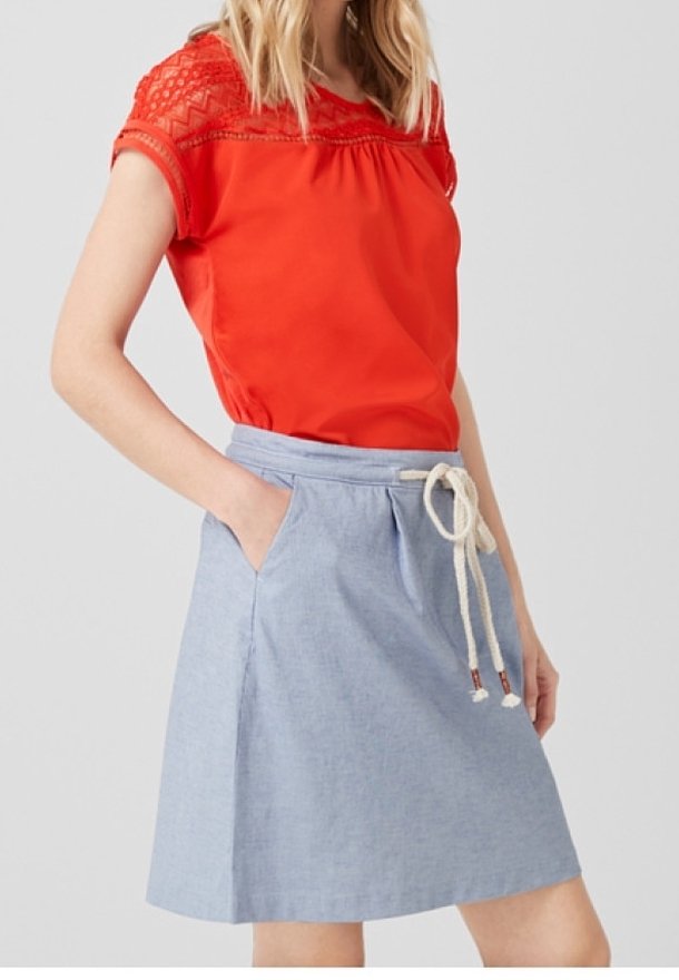 Femme portant un chemisier en dentelle rouge à manches courtes et une jupe bleu clair longueur genou avec une ceinture en corde blanche et des poches.