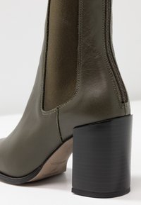 Oliwkowe kozaki sięgające kostki, wykonane ze skóry i tkaniny, z masywnym czarnym obcasem i detalami zamka błyskawicznego z boku. Gładka faktura z akcentami w postaci przeszyć.