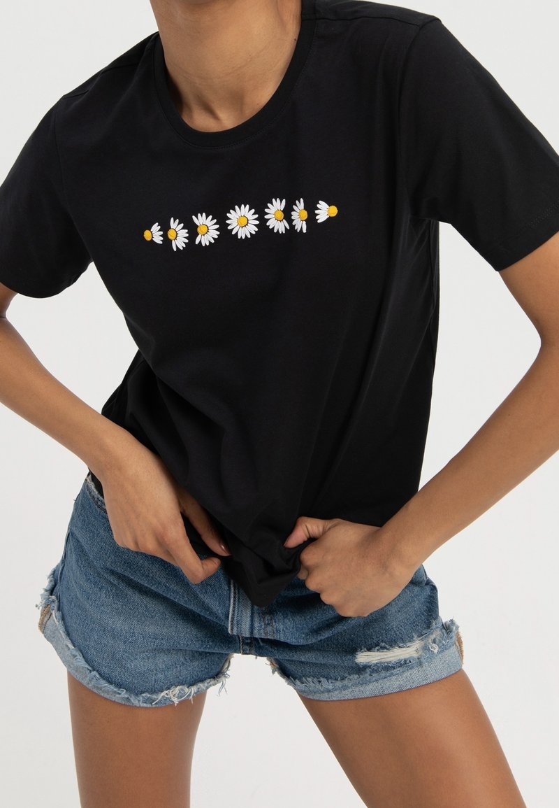 Personne portant un t-shirt noir avec une rangée de sept marguerites blanches et un short en jean bleu usé, mains ajustant l'ourlet du t-shirt.