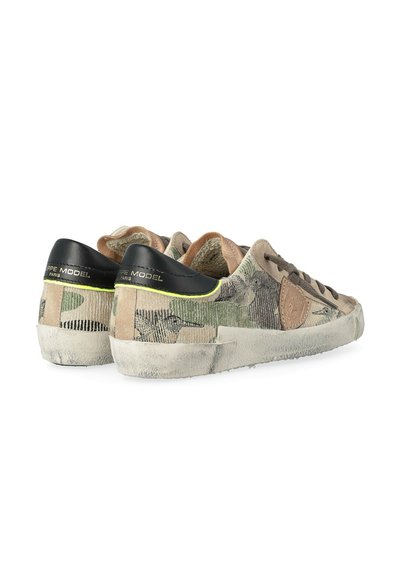 Baskets basses avec un motif camouflage beige et vert, onglets de talon noirs, semelles blanches usées et emplacements de logo en cuir marron sur les côtés.