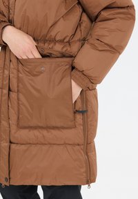 Brun puffjacka med en texturerad yta, som har en stor fyrkantig ficka och en justerbar dragkedja på sidan för en skräddarsydd passform.