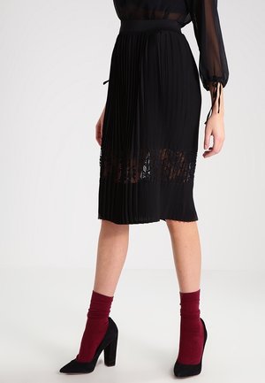 Jupe midi plissée noire avec panneau en dentelle, associée à un chemisier noir transparent, des chaussettes bordeaux et des chaussures à talons noires sur un mannequin.