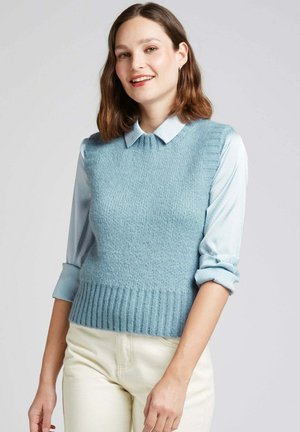 Pull sans manches en tricot bleu clair avec une taille côtelée, porté sur une chemise à col bleu clair. Texture lisse avec un motif subtil.