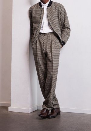 Person trägt beige-schwarz kariertes Jackett und Hose, weißes Hemd und braune Anzugschuhe, steht mit Händen in den Taschen.