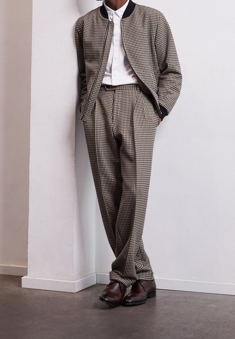 Personne portant une veste et un pantalon à carreaux beige et noir, une chemise blanche et des chaussures habillées marron, debout les mains dans les poches.