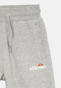 Svetlošedé tepláky s rebrovaným pásom, stiahnutím na šnúrku, bočnou kapsou a bielym logom "ellesse" s oranžovými a červenými detailmi.