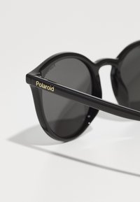 Ochelari de soare rotunzi negri cu lentile închise, având un cadru lucios și un logo auriu "Polaroid" pe tâmplă. Textură netedă, brațe groase.