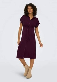 Fioletowa sukienka z guzikami, z krótkim rękawem, z paskiem w talii, sięgająca do połowy łydki. Noszona z beżowymi botkami za kostkę.