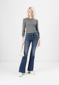 Gray long-sleeve top paired with dark blue flared jeans. Model holds a small white handbag; stands on a plain white background.