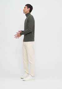 Maglione a collo alto verde oliva con un design testurizzato, abbinato a pantaloni beige chiaro e sneaker bianche, in piedi contro uno sfondo semplice.