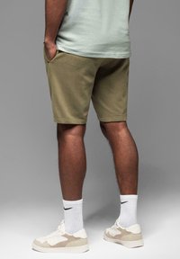 Olivengrønne shorts med en glat tekstur, sidelommer og en hældt finish, parret med cremefarvede sneakers og hvide sokker.
