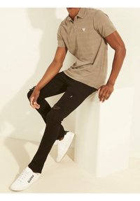 Polo kaki avec un petit logo, associé à un jean noir déchiré et des baskets blanches. La pose est assise sur un cube blanc.