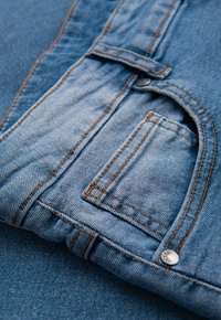 Tissu en denim bleu clair avec coutures visibles. Comprend une petite poche aux bords arrondis et un bouton argenté en guise d'accent. Texture douce.