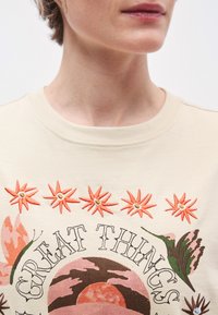 Felpa beige chiaro con fiori arancioni ricamati e testo decorativo intorno a un disegno grafico. Tessuto morbido in cotone, girocollo.