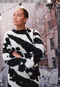 Neulepaita, jossa on mustavalkea lehmänkuvio, korkealla kaula-aukolla ja leveillä hihoilla. Pehmeä, rakenteellinen kangas näyttää lämpimältä ja mukavalta.