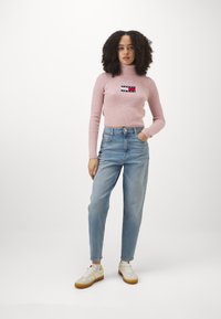 Svetlo roza rebrasti pulover s turtleneck ovratnikom in vezenim logotipom, v kombinaciji z visokim pasom svetlo modrih kavbojk in belimi supergami z rumenimi detajli.