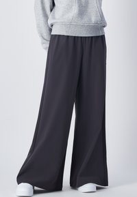 Weite Hose in Dunkelgrau, aus leichtem Stoff. Verfügt über eine glatte Textur und seitliche Taschen. Kombiniert mit einem grauen Hoodie und weißen Sneakers.