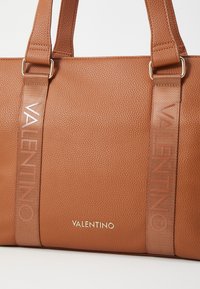 Kožená taška hnědé barvy s texturovaným povrchem, se širšími popruhy vyraženými s nápisem "VALENTINO" a zlatými akcenty loga.