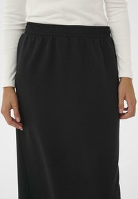 Femme portant une jupe noire à taille élastique et un haut à manches longues blanc, les mains détendues sur les côtés, devant un fond uni.