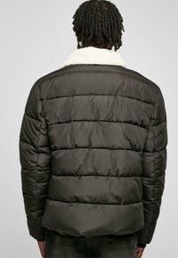 Puffjakke i mørkegrøn med horisontalt kviltet mønster, der har en cremefarvet faux shearling krave og en lynlås foran.