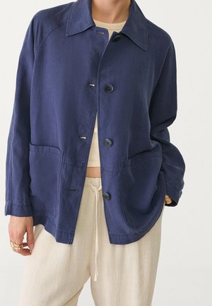 Personne portant une veste ample bleu marine boutonnée avec de grandes poches avant sur un crop top beige et un pantalon crème à cordon.