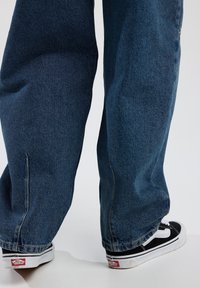 Valkene blue denim-farkut, joissa on karhea pinta ja raaka helma, yhdistetty mustiin ja valkoisiin Vans-lenkkareihin, joissa on logo.