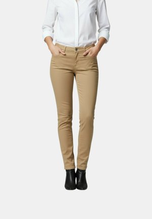 Donna con pantaloni beige aderenti, camicia bianca abbottonata con maniche arrotolate e stivaletti neri, in piedi con le mani in tasca.