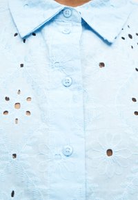 Ανοιχτό γαλάζιο πουκάμισο με κεντημένα λουλούδια eyelet, υφή σαν δαντέλα και κουμπιά στο ίδιο χρώμα μπροστά κάτω από γιακά με μυτερή άκρη.