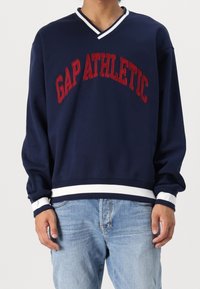 Granatowa bluza z dekoltem w serek, z czerwonym napisem "GAP ATHLETIC", białe paski na mankietach i obszyciu, wykonana z miękkiego materiału, o luźnym kroju.