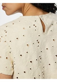 Gros plan d'une personne portant un haut brodé couleur crème avec de petits trous découpés et une encolure en forme de goutte au dos.