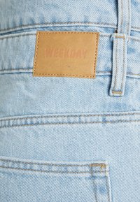 Os jeans de ganga azul claros apresentam um rótulo de couro marrom claro com a palavra "WEEKDAY". A costura visível e um bolso traseiro acentuam o design.
