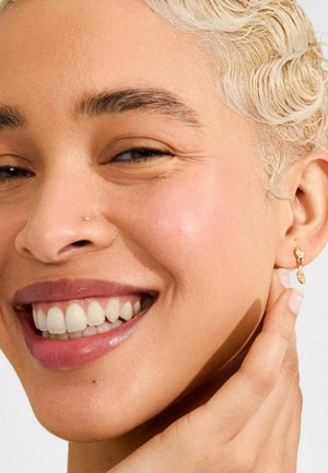 Gold earrings with a unique abstract shape, featuring a small sparkling accent, worn by a person with short, light hair.