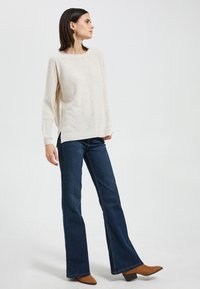 Lichtbeige gebreide trui met brede halslijn, gecombineerd met donkerblauwe flared jeans en bruine enkellaarsjes met hak. Eenvoudig ontwerp en gestructureerde stof.