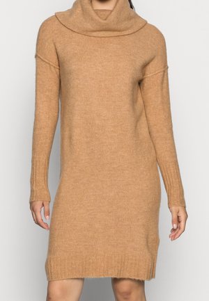 Frau in einem knielangen, haselnussbraunem Strickkleid mit langen Ärmeln und Kapuze vor einem einfarbigen Hintergrund.