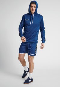 Blauwe sportieve hoodie met trekkoorden, bijpassende shorts, en zwart-witte chevron-accenten op de mouwen. Draag met donkere sneakers en witte sokken.