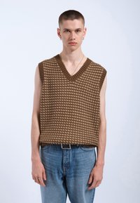 Bruine gebreide vest met een V-hals en een gestructureerd patroon, gecombineerd met lichtblauwe spijkerbroek en een zwarte riem.